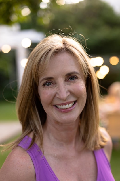 Foto retrato fechado de uma mulher caucasiana com cabelo loiro na altura dos ombros no jardim, sorrindo para a câmera, sua família sentada à mesa para uma refeição ao fundo