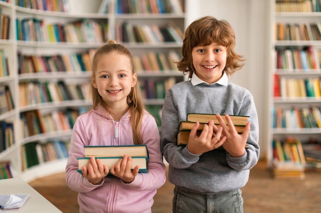 Retrato de felices alumnos europeos de niños y niñas sosteniendo una pila de libros de pie en la biblioteca y