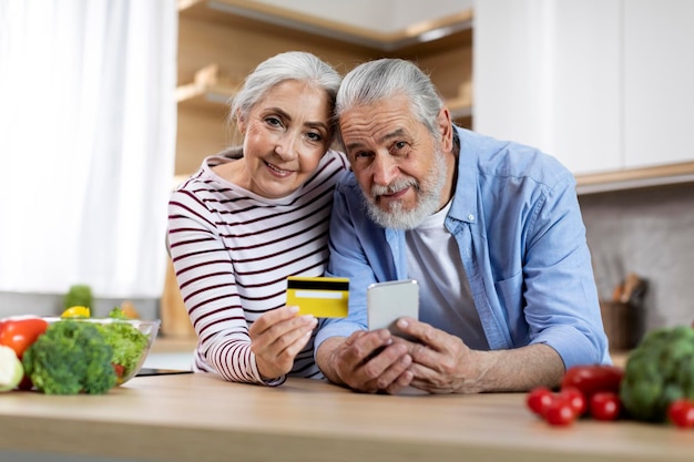 Retrato de felices cónyuges mayores con smartphone y tarjeta de crédito en la cocina