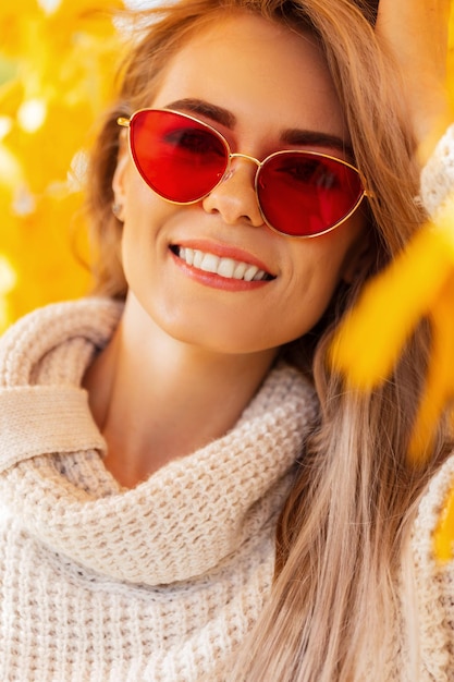 Retrato de felicidad otoñal de una hermosa joven con una sonrisa y gafas de sol rojas usando un suéter de punto de moda en las coloridas caminatas de hojas en el parque de otoño