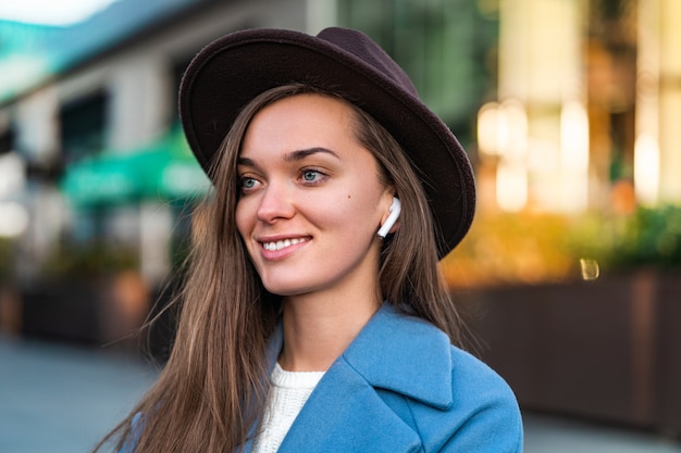 Retrato de feliz alegre elegante moda moda hipster chica con sombrero con auriculares inalámbricos blancos disfruta de la música en el centro de la ciudad. Estilo de vida y tecnología de los pueblos modernos