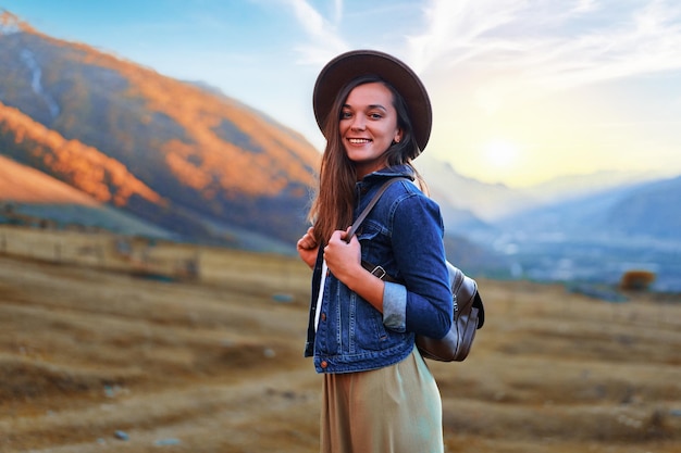 Retrato de feliz alegre joven linda sonriente hipster chica vagabundo con sombrero y mochila viajando solo en el valle de la montaña en el país de Georgia