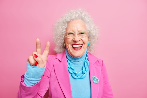 Retrato de feliz anciana europea con cabello rizado hace gesto de paz se divierte usa maquillaje brillante vestida con ropa de moda