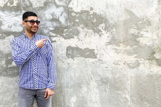 Retrato de feliz apuesto joven barbudo con camisa azul a cuadros y gafas de sol de pie contra la pared gris de hormigón. apuntando al fondo copyspace vacío y mirando a la cámara con una gran sonrisa.