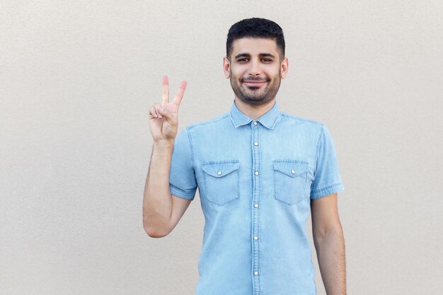 Retrato de un feliz y apuesto joven barbudo con camisa azul de pie con la victoria, o las manos de un gesto de paz y mirando la cámara y sonriendo. tiro de estudio interior aislado sobre fondo de pared beige claro.