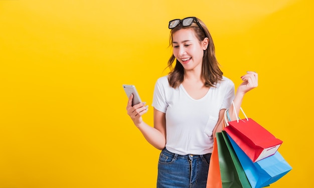 Retrato feliz asiática tailandesa hermosa mujer joven sonrisa dientes blancos soporte camiseta, ella sostiene bolsas de compras y usa la aplicación en el teléfono móvil, foto de estudio aislada en fondo amarillo con espacio de copia