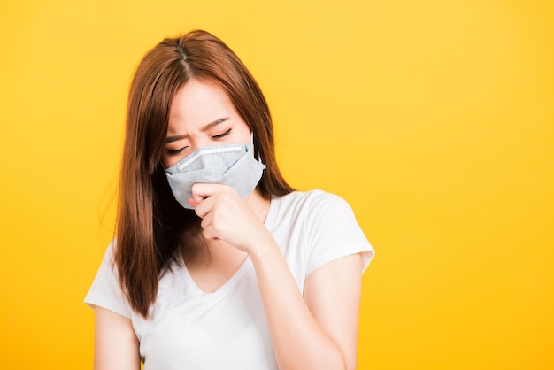 Retrato feliz asiático linda jovem adolescente em pé vestindo camiseta tosse em máscara de proteção contra epidemia de vírus ou poluição do ar isolada, tiro em estúdio em fundo amarelo com espaço de cópia