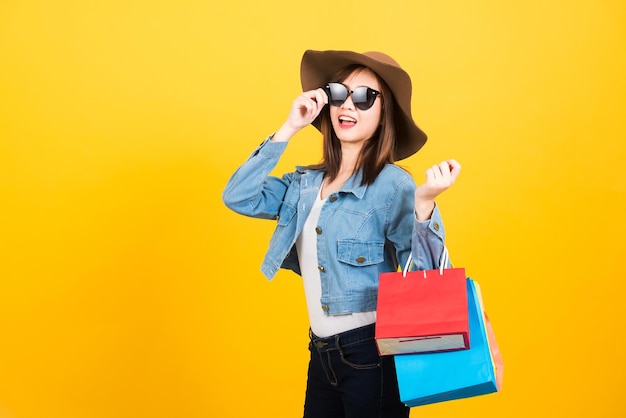 Retrato feliz asiático linda jovem adolescente sorridente em pé com óculos de sol animado segurando sacolas de compras multi cor olhando câmera isolada, estúdio tiro fundo amarelo com espaço de cópia