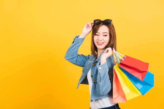 Retrato feliz asiático linda jovem adolescente sorridente em pé com óculos de sol animado segurando sacolas de compras sacolas multicoloridas isoladas, fundo amarelo com espaço de cópia