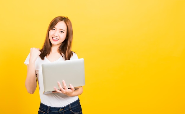 Retrato feliz asiático linda jovem adolescente sorridente em pé vestindo camiseta segurando computador portátil e animado comemorando o sucesso olhando para a câmera, fundo amarelo com espaço de cópia