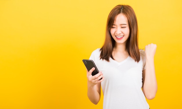 Retrato feliz asiático linda jovem adolescente sorridente vestindo camiseta comemorando a vitória com telefone móvel inteligente olhando o telefone isolado, estúdio filmado em fundo amarelo com espaço de cópia