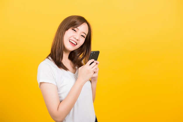 Retrato feliz asiático linda jovem adolescente sorridente vestindo camiseta de pé usando telefone celular inteligente olhando para câmera isolada, estúdio filmado em fundo amarelo com espaço de cópia