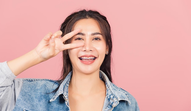 Retrato feliz asiático linda jovem bonita vestindo denim sorriso em pé mostrando dedo fazendo sinal v símbolo de vitória perto do olho olhando para câmera tiro em estúdio isolado em fundo rosa com espaço de cópia