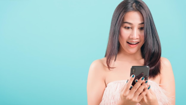 Retrato feliz asiático linda jovem em pé sorria segurando smartphone ou celular e olhando para o telefone sobre fundo azul com espaço de cópia para texto