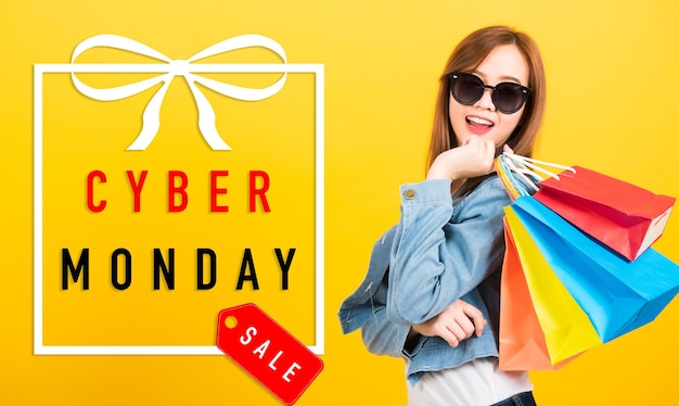 Retrato feliz asiático linda jovem sorrindo com óculos de sol animado segurando sacolas de compras câmera multicolorida com texto Cyber Monday na caixa de presente no fundo amarelo isolado lateral