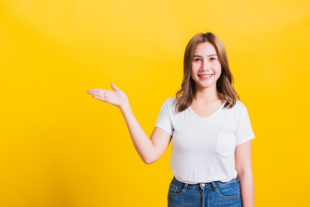 Retrato feliz asiático tailandês linda jovem bonita em pé vestindo camiseta branca segurando algo na palma da mão olhando para a câmera, tiro de estúdio isolado em fundo amarelo com espaço de cópia