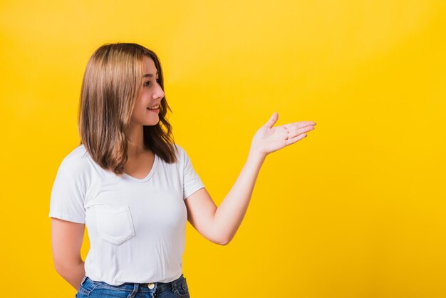 Retrato feliz asiático tailandês linda jovem bonita em pé vestindo camiseta branca segurando algo na palma da mão olhando para o lado, tiro de estúdio isolado em fundo amarelo com espaço de cópia