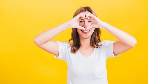 Retrato feliz asiático tailandês linda jovem bonita sorriso em pé faça o dedo coração figura símbolo forma sinal com as duas mãos e olhando a câmera, estúdio tiro isolado fundo amarelo com espaço de cópia