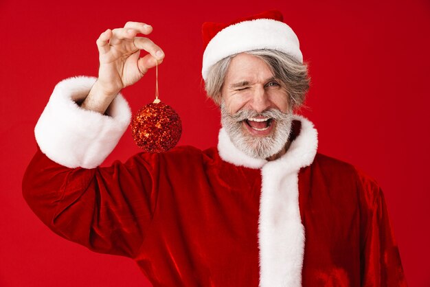 Retrato de feliz canoso viejo hombre de Santa Claus sonriendo y sosteniendo la bola del árbol de Navidad aislado en rojo en estudio