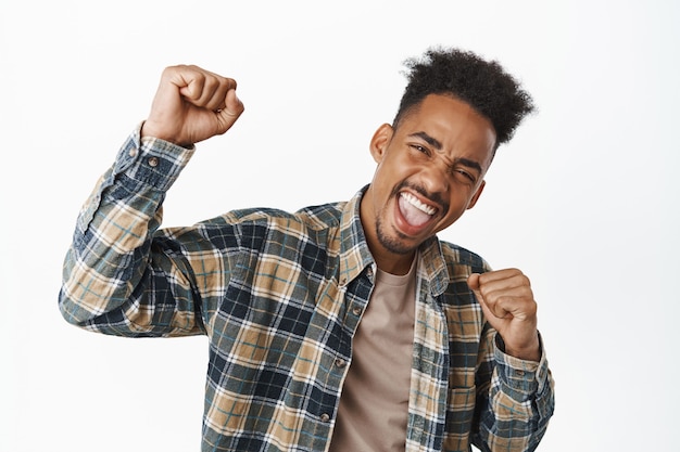 Retrato de feliz chico afroamericano cantando, levantando el puño y gritando de regocijo, emocionado por ganar, celebrando la victoria, ganando el juego, de pie con camisa a cuadros en blanco