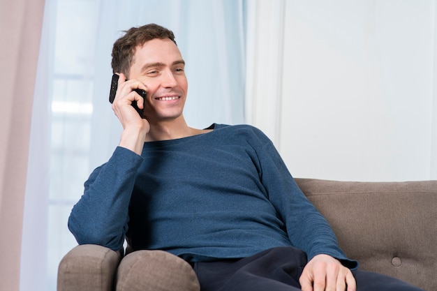 Retrato de feliz chico positivo, joven alegre está charlando, hablando por teléfono celular, teniendo una buena conversación agradable en el teléfono inteligente, sentado en el sofá o el sofá en la sala de estar en casa, sonriendo