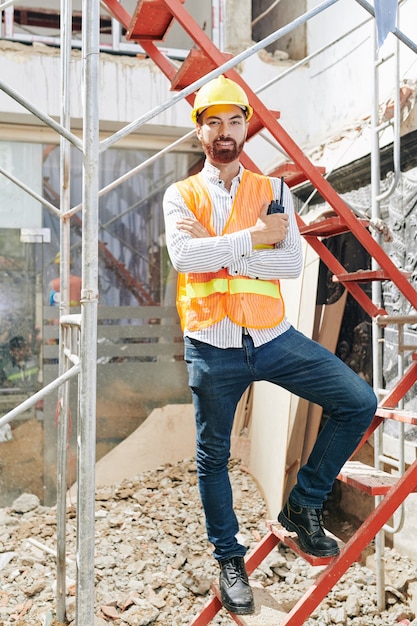 Retrato de feliz contratista confiado en chaleco naranja brillante y casco de pie en la escalera en edificio en construcción con walkie-talkie en manos