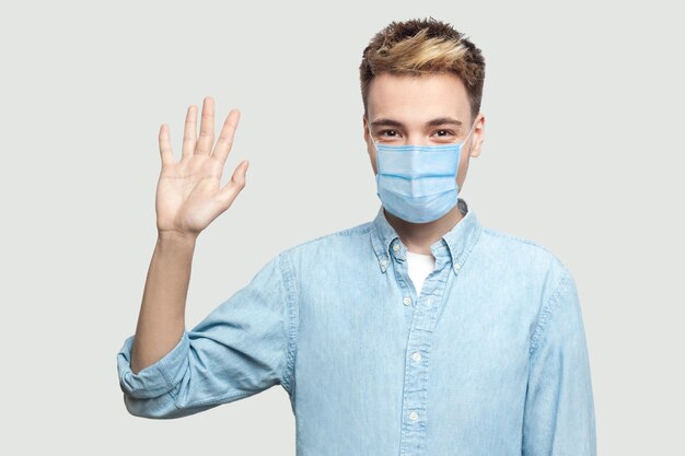 Retrato de feliz emocionado apuesto joven con máscara médica quirúrgica en camisa azul de pie, mirando a cámara y agitando su mano para saludar. tiro del estudio de interior en el espacio gris de la copia del fondo.