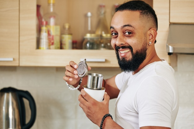 Retrato de feliz emocionado joven barbudo de pie en la cocina con gotero de café de aluminio