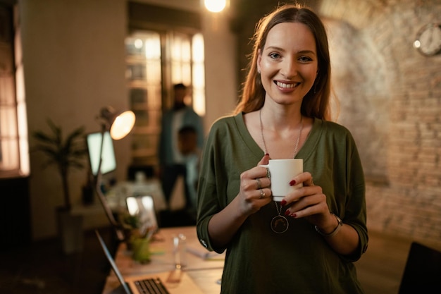 Retrato de feliz empresaria creativa con taza de café por la noche en la oficina