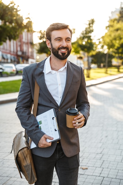 Retrato de feliz empresario adulto en traje formal sosteniendo café para llevar y gráficos de papel mientras camina por las calles de la ciudad