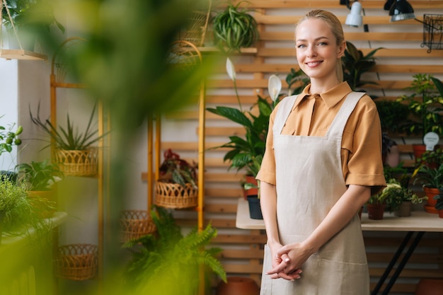 Retrato de feliz y encantadora florista femenina con delantal de pie posando en una tienda de flores mirando a la cámara