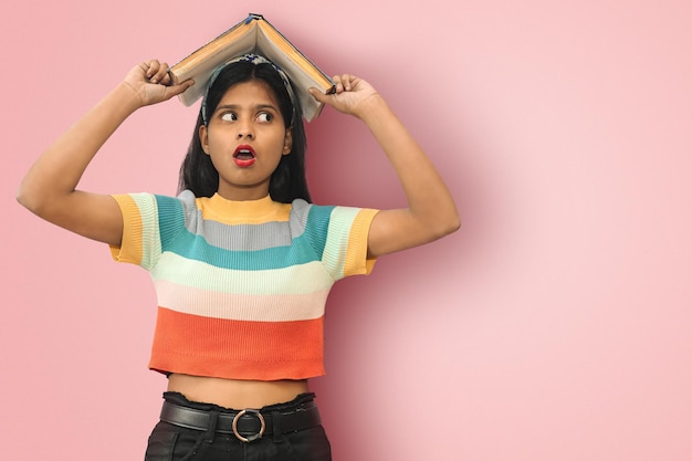 Retrato de una feliz estudiante india asiática sorprendida con la boca abierta y los ojos grandes posando aislada sosteniendo un libro en la cabeza y mirando hacia el espacio de la copia