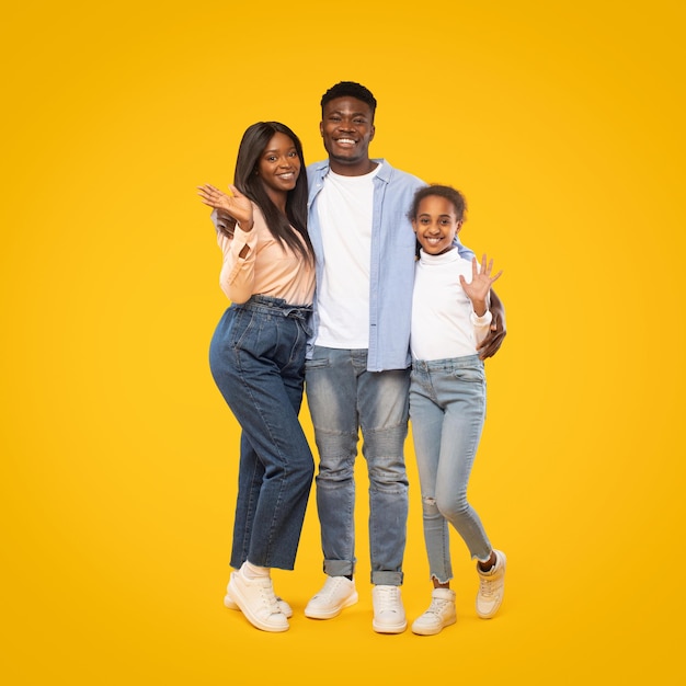 Retrato de una feliz familia afroamericana de hombres, mujeres y niñas saludando con la mano mientras posan sobre un fondo amarillo