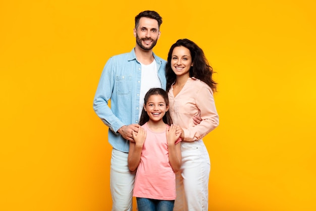 Retrato de una feliz familia europea de tres abrazándose y sonriendo a la cámara posando juntos en amarillo