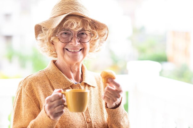 Retrato de feliz hermosa anciana rizada en amarillo con sombrero y gafas de sol sosteniendo una taza de café y una magdalena al aire libre