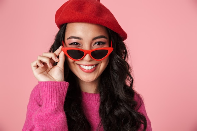 Retrato de feliz hermosa chica asiática con cabello largo y oscuro en boina con elegantes gafas de sol aisladas en rosa