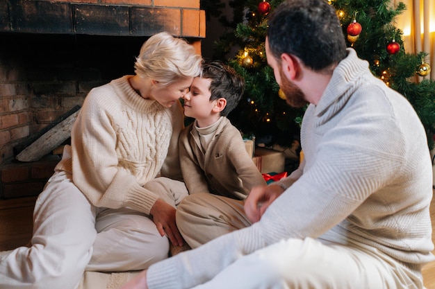 Retrato de feliz hermosa familia caucásica sentado en abrazos por árbol de Navidad y acogedora chimenea