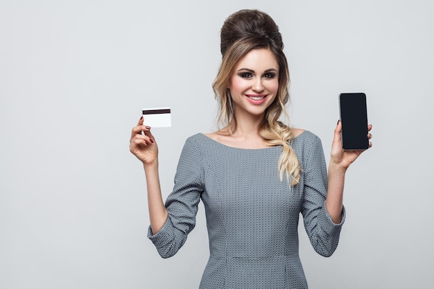 Retrato de feliz hermosa joven moderna en vestido gris de pie, sosteniendo el teléfono celular y la tarjeta de crédito con una gran sonrisa, mirando a la cámara. Interior, aislado, foto de estudio, fondo gris, espacio de copia