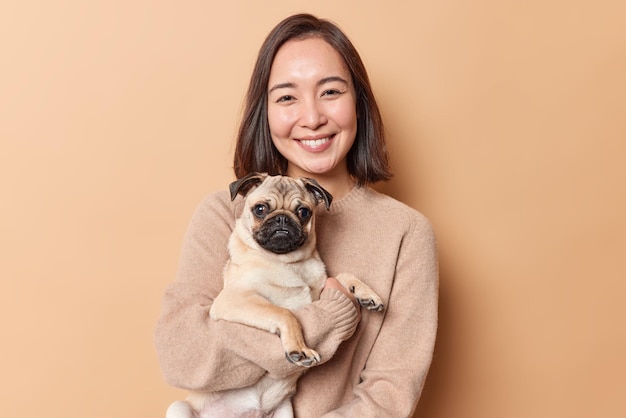 Retrato de feliz hermosa joven mujer asiática con expresión tierna y alegre contenta de tener un perro pug como regalo vestido con un puente suave aislado sobre fondo marrón. Concepto de cuidado de personas y mascotas.
