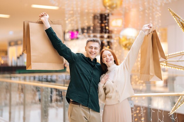 Retrato de feliz hermosa joven pareja enamorada sosteniendo bolsas de compras de pie en el centro comercial y mirando