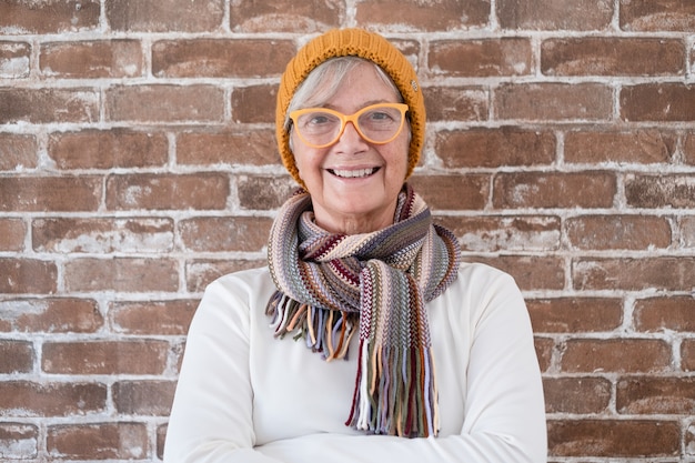 Foto retrato de feliz hermosa mujer mayor vistiendo gorro y bufanda de invierno - fondo de pared de ladrillo