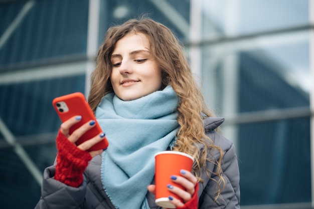 Retrato de feliz hermosa mujer rizada con taza de café para llevar usar mensajes de texto de teléfono móvil.