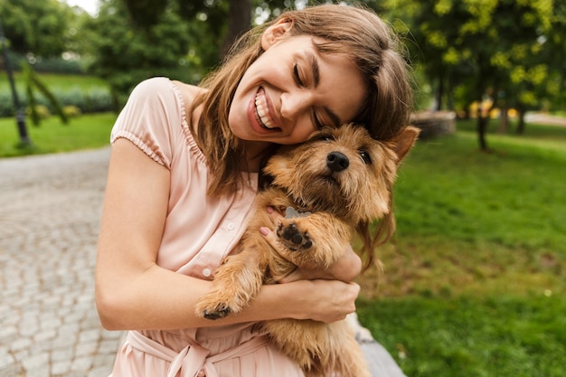 Retrato de feliz hermosa mujer vestida con vestido sonriendo y abrazando a perro mientras camina en un parque