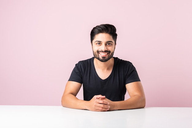 Retrato de feliz hombre barbudo asiático indio sentado en una mesa o escritorio o plataforma contra el fondo de color rosa mirando a la cámara