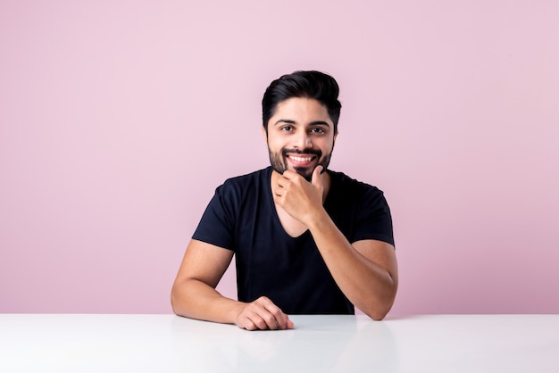 Retrato de feliz hombre barbudo asiático indio sentado en una mesa o escritorio o plataforma contra el fondo de color rosa mirando a la cámara