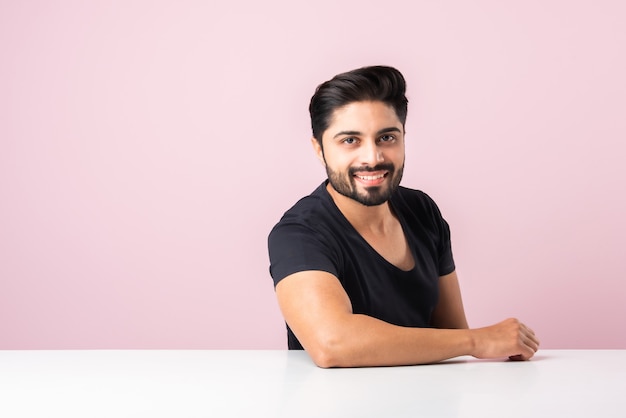 Retrato de feliz hombre barbudo asiático indio sentado en una mesa o escritorio o plataforma contra el fondo de color rosa mirando a la cámara