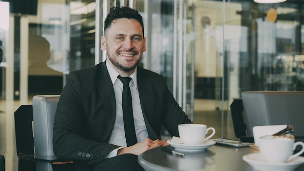 Retrato de feliz hombre de negocios caucásico barbudo en ropa formal sentado bebiendo café y smili