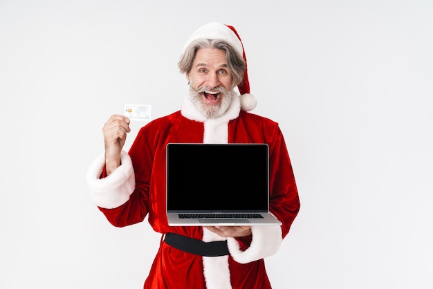 Retrato de feliz hombre de pelo gris de Santa Claus en traje rojo con portátil y tarjeta de crédito aislado en blanco