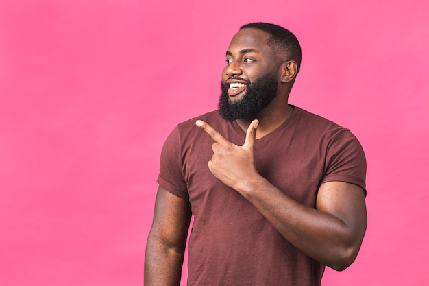 Retrato de feliz joven afroamericano negro con una sonrisa casual, apuntando a un lado con el dedo, mirando a puerta cerrada con expresión de la cara emocionada aislada sobre fondo rosa.