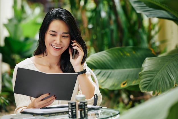 Retrato de feliz joven asiática leyendo el catálogo, hablando por teléfono y pidiendo ingredientes para su marca de cosméticos